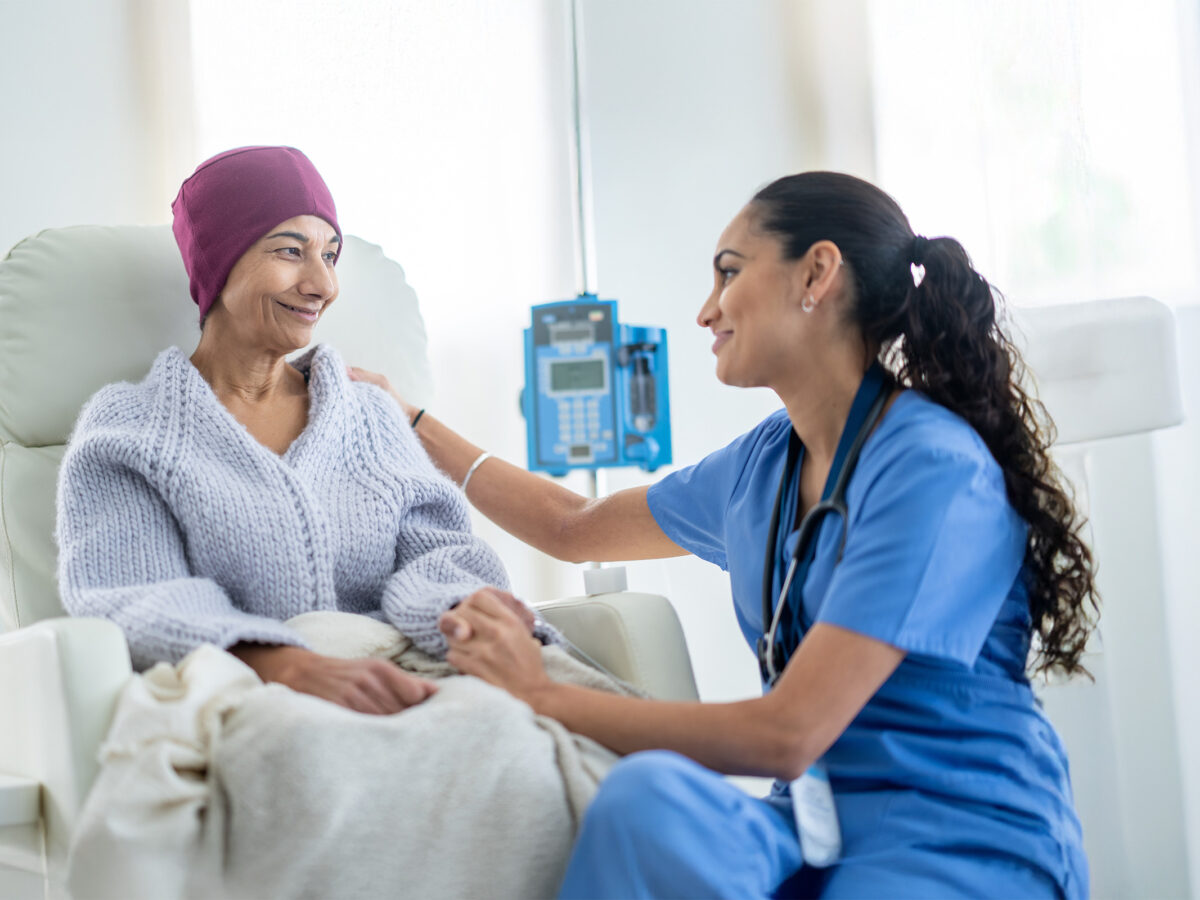 A senior woman battling cancer sits in a high back chair as she talks with her nurse during a Chemotherapy treatment. The patent is dressed casually in a sweater and has a blanket and headscarf on to keep her warm.