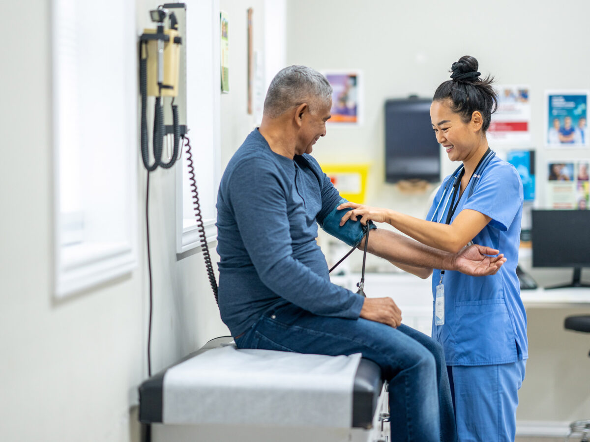 A senior gentleman of Hispanic decent, sits up on an exam table as his female nurse takes his blood pressure. The nurse is wearing blue scrubs and making small talk to put the gentleman at ease.