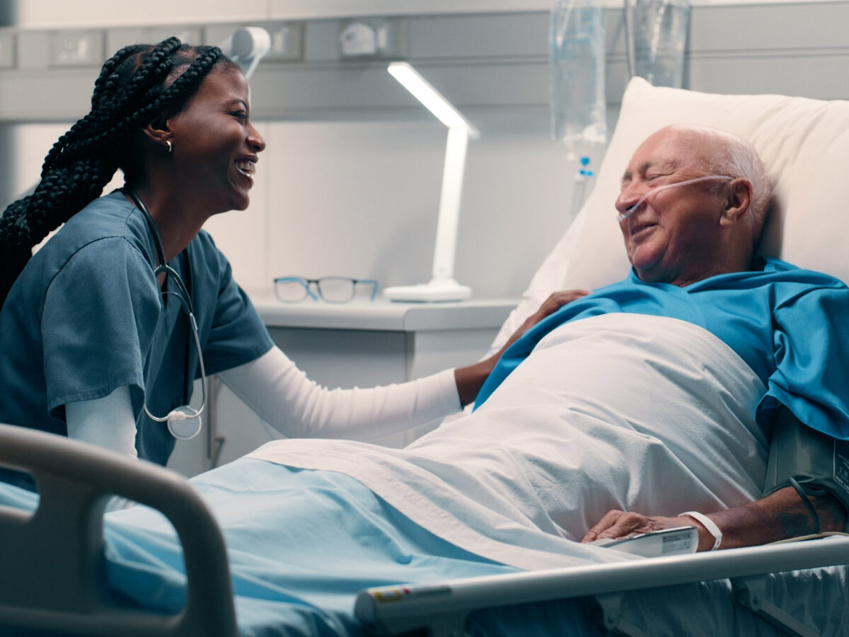 Bed, nurse and smile with patient in hospital for help, support and good news with care. People, professional and happy for healthcare for illness or sickness and recovery with treatment or medical